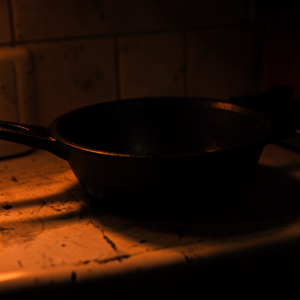 Close-up dark photograph of cast-iron cookware resting on a worn kitchen counter, lit by a single warm amber glow
