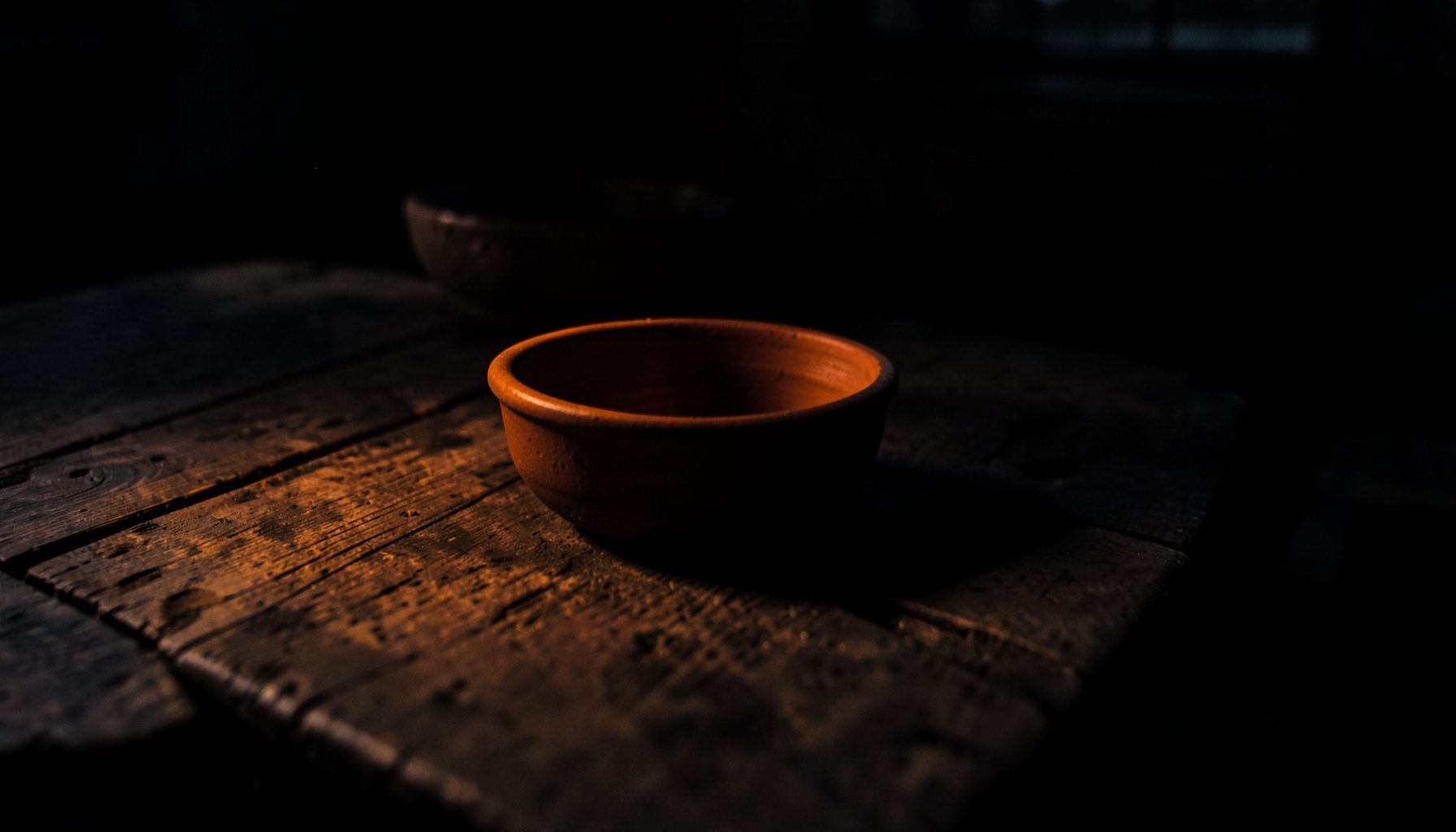 Dark, low-key photograph of a wooden farmhouse table at dusk, with deep shadow and warm amber light spilling across rough boards and a clay bowl, conveying a rustic, moody atmosphere in deep charcoal and ember tones