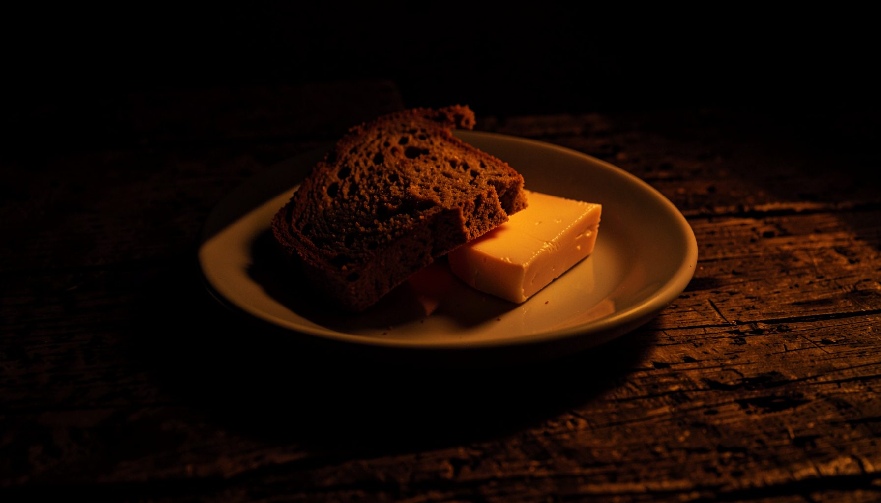 Atmospheric dark photograph of a simple plate of bread and cheese on a rough timber surface, lit by warm amber light against deep shadow