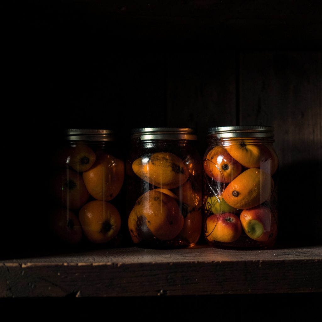 Low-key still life of amber glass jars filled with preserved fruit, sitting on a dark timber shelf with soft, deep shadow