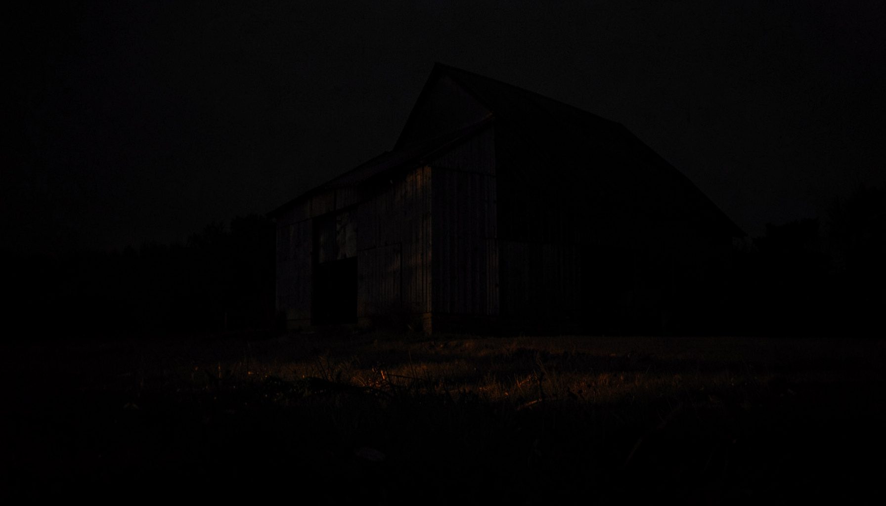 Dark, low-key photograph of a weathered barn at the edge of a field, almost lost in deep charcoal shadow with a faint amber rim of light