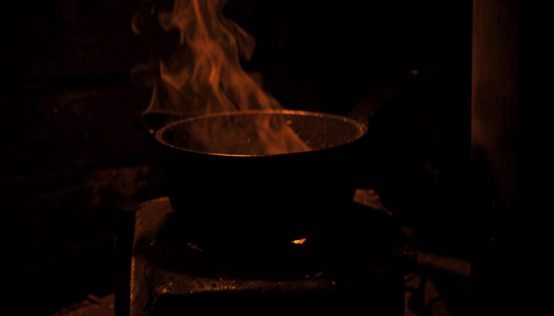 Dark still life of a steaming cast-iron pot on a woodstove, deep shadow with ember-orange light