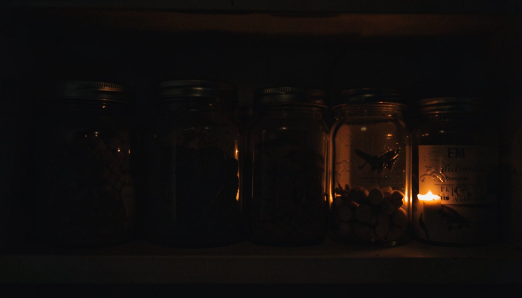 Muted, low-key photograph of a row of glass jars on a pantry shelf, lit by amber candlelight against deep shadow