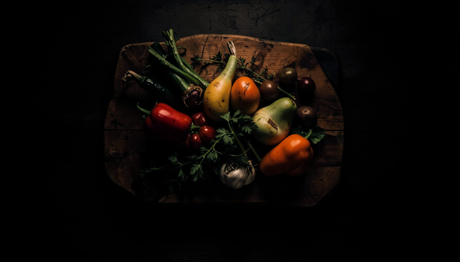 Dark overhead photograph of a rustic wooden board holding seasonal vegetables, cast in deep charcoal shadow with amber highlights, moody and atmospheric