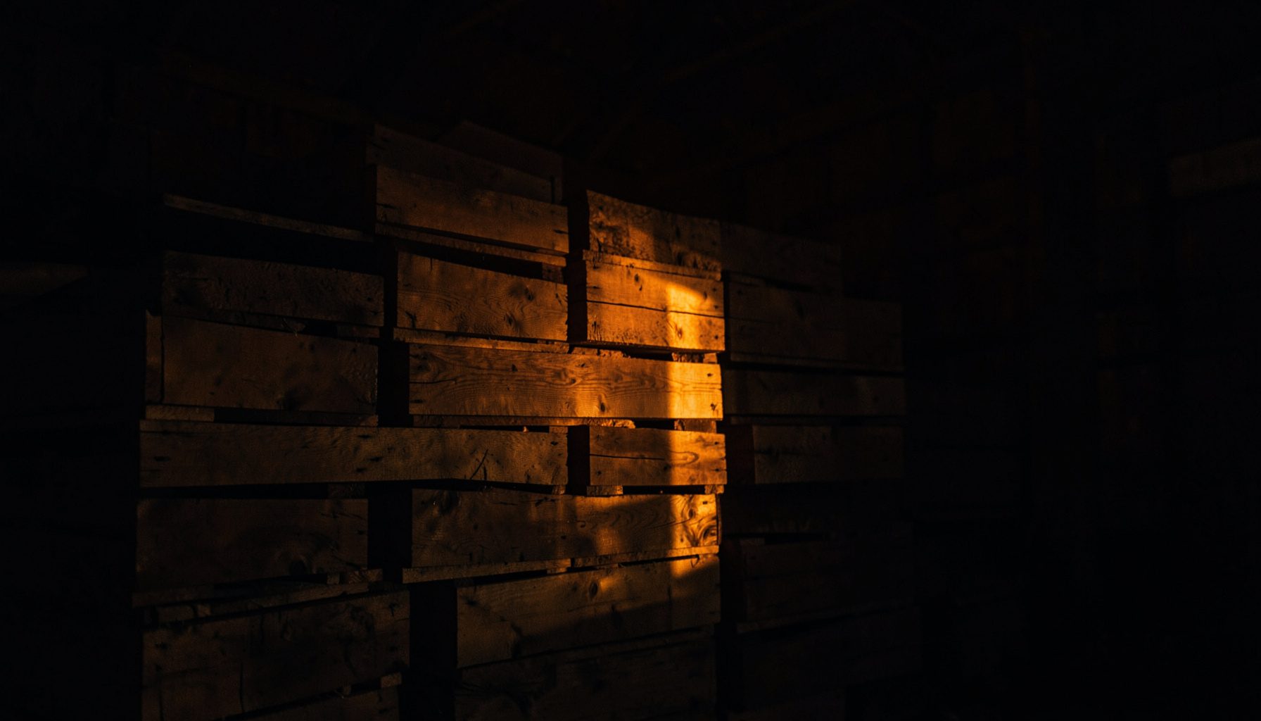 Low-key photograph of stacked wooden crates in a dim barn interior, lit by a single amber shaft